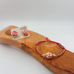 May include: A set of floral jewelry, including stud earrings and a bracelet. The earrings are on a small card, featuring a red and white flower design. The bracelet has a red cord and gold chain with a matching floral bead.