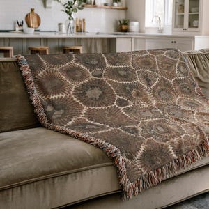 May include: A brown and beige throw blanket with a repeating circular pattern, fringed edges, and a soft texture. The blanket is draped over a brown sofa in a well-lit living room setting.