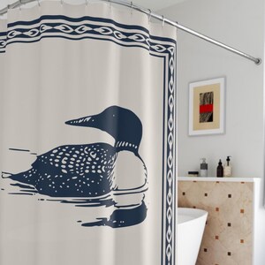 A white shower curtain featuring a navy blue illustration of a loon swimming on water. The loon is detailed with white spots and a black head. The curtain has a navy blue border with a decorative pattern, and is hanging in a rustic bathroom.