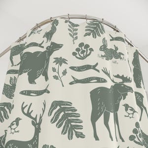 A shower curtain with a repeating pattern of forest animals in dark green on a cream background. The woodland design includes bears, deer, rabbits, foxes, birds, fish, and plants. The curtain has a whimsical log cabin aesthetic.