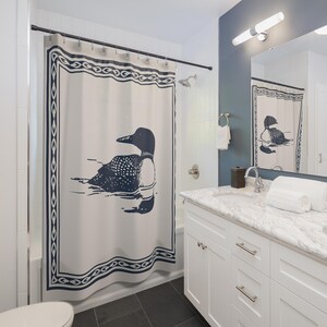 A white shower curtain featuring a navy blue illustration of a loon swimming on water. The loon is detailed with white spots and a black head. The curtain has a navy blue border with a decorative pattern, and is hanging in a rustic bathroom.
