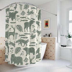 A shower curtain with a repeating pattern of forest animals in dark green on a cream background. The woodland design includes bears, deer, rabbits, foxes, birds, fish, and plants. The curtain has a whimsical log cabin aesthetic.