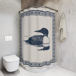 A white shower curtain featuring a navy blue illustration of a loon swimming on water. The loon is detailed with white spots and a black head. The curtain has a navy blue border with a decorative pattern, and is hanging in a rustic bathroom.