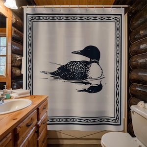 A white shower curtain featuring a navy blue illustration of a loon swimming on water. The loon is detailed with white spots and a black head. The curtain has a navy blue border with a decorative pattern, and is hanging in a rustic bathroom.