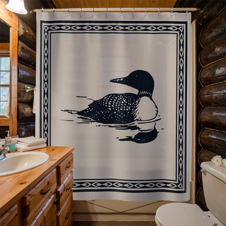 A white shower curtain featuring a navy blue illustration of a loon swimming on water. The loon is detailed with white spots and a black head. The curtain has a navy blue border with a decorative pattern, and is hanging in a rustic bathroom.