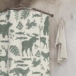 A shower curtain with a repeating pattern of forest animals in dark green on a cream background. The woodland design includes bears, deer, rabbits, foxes, birds, fish, and plants. The curtain has a whimsical log cabin aesthetic.