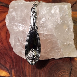 Puede incluir: Un collar con colgante plateado hecho con una cuchara. El colgante tiene un diseño floral detallado en el mango y un cuenco decorativo. El collar se exhibe sobre una gran roca blanca similar a un cristal.
