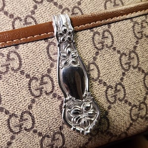 May include: A silver spoon pendant with floral detailing. The pendant is attached to a brown and tan patterned handbag. The spoon's design features detailed flower and leaf patterns.