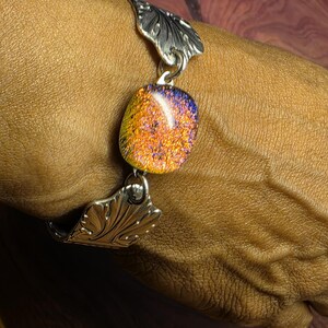 Puede incluir: Una pulsera que presenta un cabujón central de vidrio dicroico rectangular con tonos brillantes en naranja, amarillo y morado, flanqueado por dos eslabones de metal plateado con un patrón de hojas en relieve y unidos por anillas.