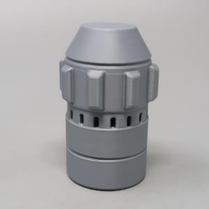 Puede incluir: Objeto cil&iacute;ndrico gris con una parte superior c&oacute;nica. Presenta protuberancias rectangulares y peque&ntilde;os agujeros espaciados uniformemente alrededor de la secci&oacute;n media. La base es un cilindro m&aacute;s ancho. Parece ser impreso en 3D.