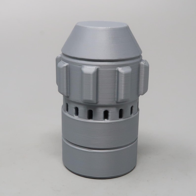 Puede incluir: Objeto cil&iacute;ndrico gris con una parte superior c&oacute;nica. Presenta protuberancias rectangulares y peque&ntilde;os agujeros espaciados uniformemente alrededor de la secci&oacute;n media. La base es un cilindro m&aacute;s ancho. Parece ser impreso en 3D.