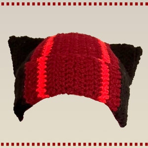 Könnte beinhalten: Eine rote und schwarze Häkelmütze mit Katzenohren. Die Mütze hat rote Streifen und schwarze Ohren.
