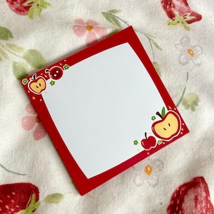 Puede incluir: Un bloc de notas cuadrado con borde rojo e interior blanco. Las esquinas están decoradas con ilustraciones de manzanas de dibujos animados. Descansa sobre una tela floral con fresas y flores.