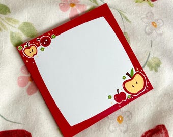 May include: A square notepad with a red border and a white interior. The corners are decorated with cartoon apple illustrations. It sits on floral fabric with strawberries and flowers.