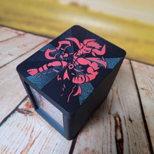 May include: A black wooden box with a red graphic of a warrior figure on the top. The graphic is set against a dark blue, starburst-like background. The box has a clear window on one side.