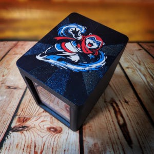 May include: A black box with a clear window and a cartoon graphic on top. The graphic features a character in a red scarf and goggles, with blue and white accents. The box is on a wooden surface.