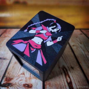 Pode incluir: Uma caixa retangular preta com uma ilustração gráfica de uma personagem com cabelo trançado, vestindo um traje rosa e roxo. O design inclui um fundo escuro e brilhante com o texto AQUESTIX.COM impresso no canto inferior.