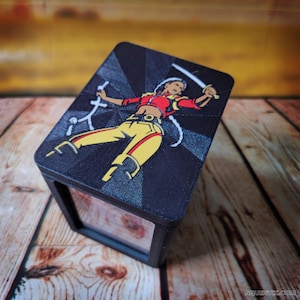 May include: A black box with a colorful illustration of a female warrior on top. The warrior is depicted in a dynamic pose, holding a sword and a small weapon. She is wearing a red top, yellow pants, and a yellow belt. The background is a dark gray with radial lines.