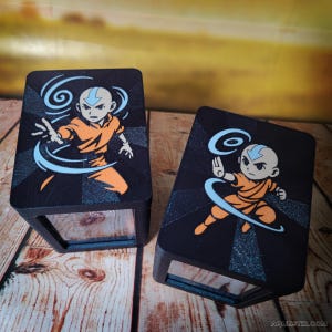 Peut inclure: Deux présentoirs rectangulaires noirs avec un personnage de dessin animé en tenue orange et détails bleus. Le personnage a la tête chauve et une flèche bleue sur le front, entouré d'énergie bleue tourbillonnante. Sur une surface en bois.