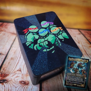 May include: A black card holder featuring a graphic of the Teenage Mutant Ninja Turtles in green, with red, blue, purple, and orange headbands. A trading card with the text "Heroes in a Half Shell" is visible.