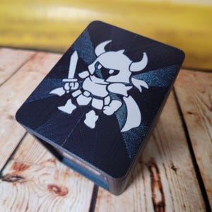 May include: A rectangular, dark blue box with a white illustration of a knight holding a sword. The knight is wearing armor and a helmet with horns. The box has a textured surface and is sitting on a wooden surface.