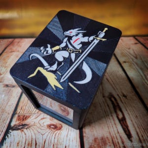 May include: A black rectangular box with a lid featuring a graphic of a white dragon-like creature holding a sword. The creature has red eyes and is positioned against a black and dark gray sunburst pattern with a yellow lightning bolt shape below.