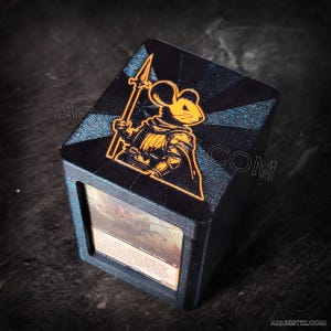 Puede incluir: Una caja de mazo negra y azul con un diseño de caballero ratón amarillo. La caja tiene una ventana transparente para mostrar una carta con el texto "Criatura legendaria - Caballero ratón".