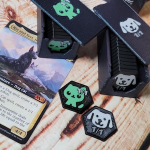 Pode incluir: Suportes de fichas pretos com desenhos de gato verde e cão branco. Os suportes estão abertos, revelando pilhas de fichas. Dois tokens hexagonais com um gato verde e um cão branco são visíveis. Uma carta de Magic: The Gathering também está na imagem.