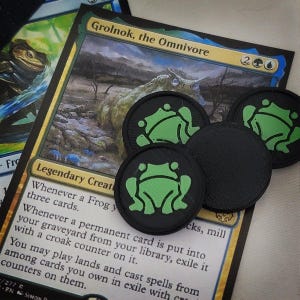 May include: Four black and green frog counters for a board game. The counters are round and have a raised frog design. The counters are on top of a Magic: The Gathering card with the text "Grolnok, the Omnivore" and game instructions.