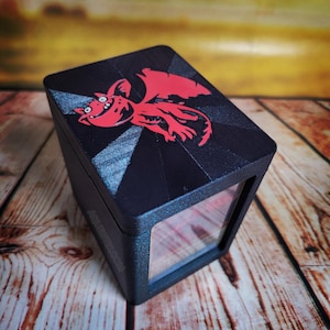 May include: Black box with a red cartoon dragon design on the top. The box has a clear window on the front. The text "AQUESTIX.COM" is visible on the side of the box. The box is sitting on a wooden surface.