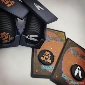 May include: Black and orange game tokens with a detective design, displayed with Magic: The Gathering cards. The tokens are in dark blue boxes. One token has a detective design, and another has a white design.