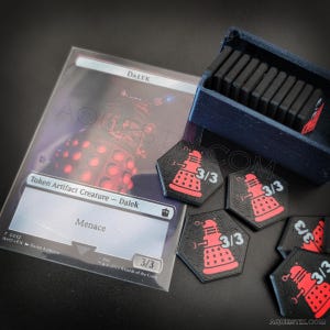 Könnte beinhalten: Eine transparente Karte mit dem Wort "DALEK" und "Token Artifact Creature - Dalek" aufgedruckt. Mehrere schwarze und rote sechseckige Token mit einem Dalek-Design und "3/3" sind sichtbar. Eine dunkelblaue Box enthält schwarze rechteckige Teile.