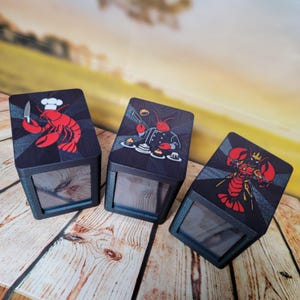 May include: Three black boxes with clear viewing windows. Each box features a different cartoon lobster design on top: one with a chef's hat and knife, one as a chef, and one wearing a crown. The lobster illustrations are red.