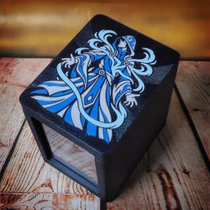 May include: A black rectangular storage box featuring a graphic illustration on the lid. The design depicts a hooded figure in a flowing blue and white robe with swirling patterns. The box has a textured matte finish and a clear front panel.