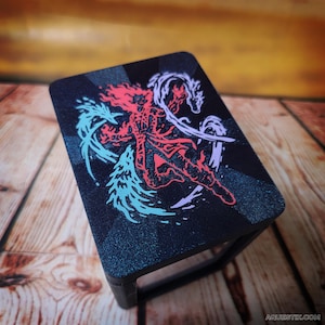 May include: A black rectangular stand with a graphic design featuring a red figure, a white dragon, and blue and white elements. The stand is on a wooden surface. The text "AQUESTIX" is visible in the bottom right corner.