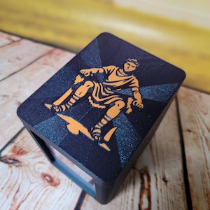 May include: A dark blue box with an orange illustration of a figure in classical attire. The figure is in motion, set against a starburst design. The box is on a wooden surface.