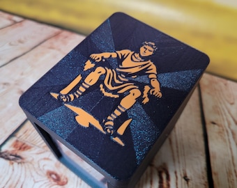 May include: A dark blue box with an orange illustration of a figure in classical attire. The figure is in motion, set against a starburst design. The box is on a wooden surface.