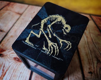 May include: Black rectangular box with a stylized, skeletal creature design in beige. The creature has a skull-like head and long, clawed hands. The text "AQUESTIX.COM" is visible on the bottom right corner.