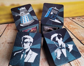 May include: Four black card storage boxes with images of characters and objects from the Doctor Who series. The boxes feature a Dalek, the TARDIS, and two portraits. The boxes are on a wooden surface.