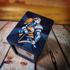 May include: A black display box with a detailed illustration of a female archer in orange, blue, and white. The archer is holding a bow and arrow. The box has a geometric pattern on the top and is set on a wooden surface.
