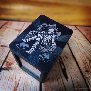 May include: A black box with a clear window and a decorative top featuring a mummy design in white and gray, surrounded by gray rats. The top has a dark blue background with a glittery effect.