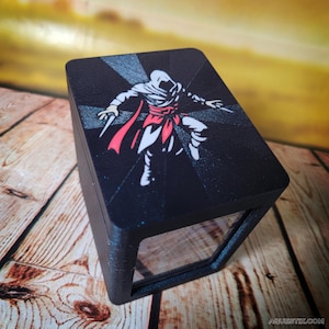 May include: Black box with a clear front panel. The top of the box features a graphic of an assassin in white, red, and black. The assassin is holding two knives. The box is sitting on a wooden surface.