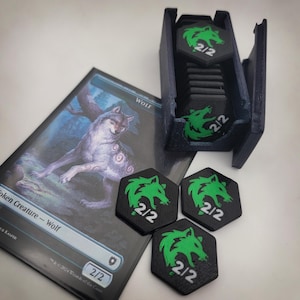 Op de afbeelding: Een Magic: The Gathering Wolf token kaart en bijpassende zeshoekige tokens met een groene wolfskop en de tekst "2/2". Een donkerblauwe tokenhouder is gevuld met vergelijkbare tokens. De kaart toont een wolf illustratie.