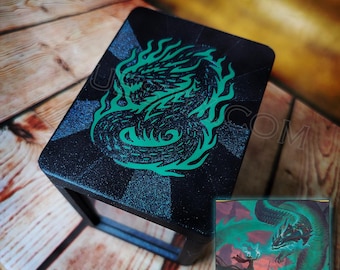 May include: Black box with a green dragon design. The dragon is surrounded by flames and is set against a dark background. The box has a rectangular shape and is sitting on a wooden surface. The text "AQUESTIX.COM" is visible.