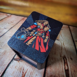 May include: A rectangular black box with a glittery finish featuring a graphic illustration of a character in a red coat and tan armor holding a blue glowing orb. The character has long hair and is depicted against a dark background with rays.