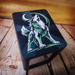 Op de afbeelding: Een zwarte, rechthoekige doos met een witte wolf die naar een maan huilt, met groene details. De achtergrond is donkerblauw met glitter. De doos heeft een zwart frame en staat op een houten oppervlak.