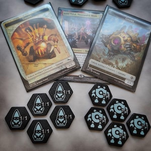 Puede incluir: Un conjunto de nueve piezas de juego hexagonales negras con un 1/1 blanco impreso en cada una. Las piezas presentan dos diseños diferentes: un gnomo y un robot. Las piezas están dispuestas en círculo sobre una mesa con tres cartas de Magic: The Gathering en el fondo. Las cartas se titulan "GNOME", "Anim Pakal, Thousandth Moon", y "GNOME".