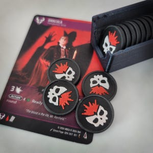 May include: A Dracula character card featuring a red and black background with the text "The blood is the life, Mr. Renfield." Next to the card are several black circular game tokens with a white skull and red crown graphic, stored in a dark holder.