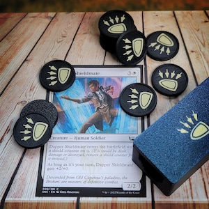 Op de afbeelding: Een Magic: The Gathering kaart met een menselijke soldaat genaamd "Dapper Shieldmate" met een mana-kost van 3 en een kracht en uithoudingsvermogen van 2/2. De kaart is omringd door zwarte en gele tokens met een pootprintontwerp.