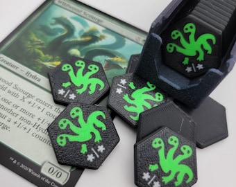 Puede incluir: Fichas de juego hexagonales negras con un diseño verde de criatura de tres cabezas y símbolos blancos, junto a una carta que dice "Wildwood Scourge". Un contenedor azul oscuro contiene fichas adicionales. Las fichas son probablemente para un juego de mesa.